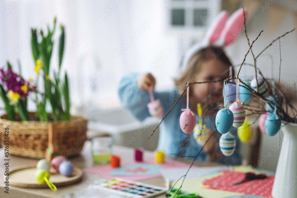 Portrait of cute pretty little girl in blue dress and funny rabbit ears making craft, diy eggs for easter holiday home decoration. Spring colours, bright flowers. Kid's domestic life, hobby, enjoyment