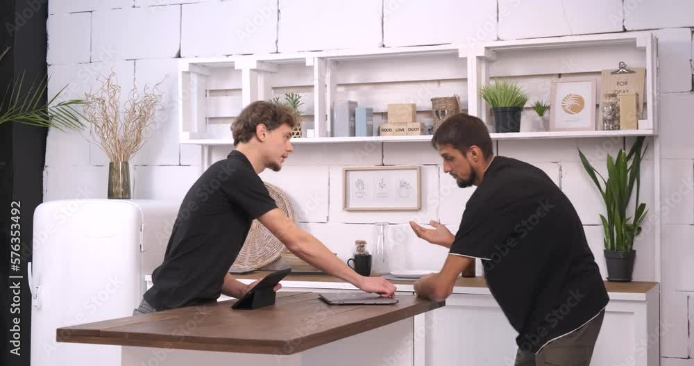 Two young men are using a digital tablet and laptop in the kitchen. Have fun spending free time together. Look at the screen and laugh Two young men are using a digital tablet and laptop in the kitchen. Have fun spending free time together. Look at the screen and laugh