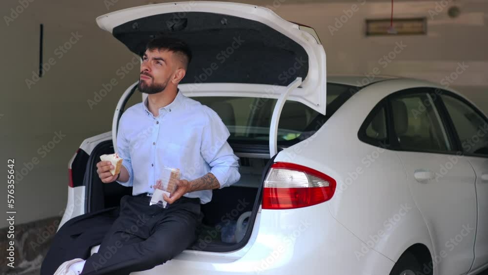 Tattooed young man eating sandwich sitting on car trunk in garage. Portrait of relaxed Caucasian guy enjoying snack indoors Tattooed young man eating sandwich sitting on car trunk in garage. Portrait of relaxed Caucasian guy enjoying snack indoors
