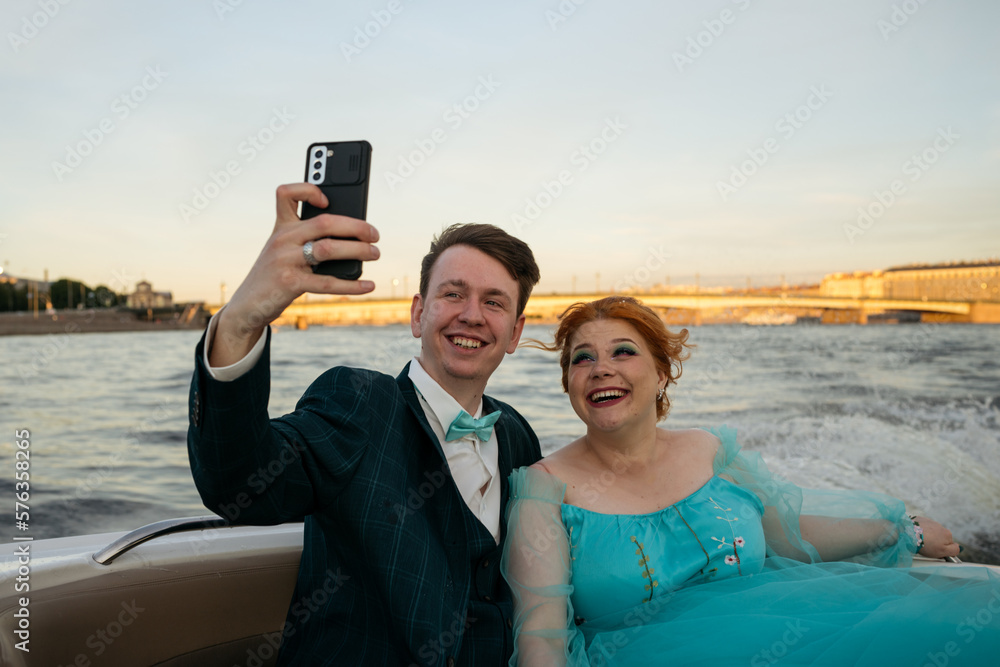 Portrait of redhead young bride in blue dress and groom in motorboat taking selfie. High speed, water splashes, sincere emotions.