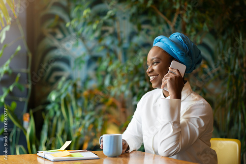 Cheerful black lady talking on phone with clients, cafe interior