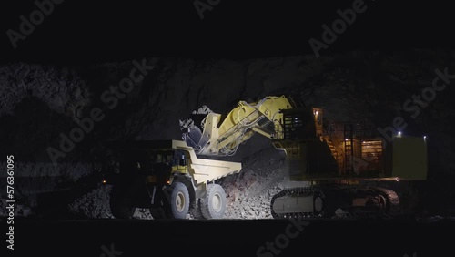 Large escavator loading ore rock in dumper truck at night, slow motion