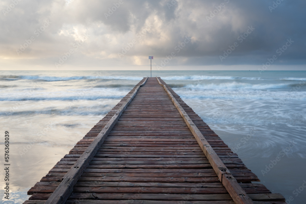 a wooden pier leading into a rough sea under a cloudy sky. long exposure