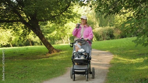 Mom and child outdoors, happy mother drinks coffee while walking in the summer park with her young daughter in a baby carriage