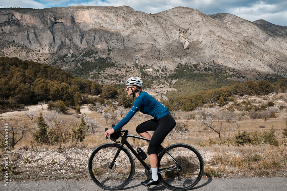 With a view of the beautiful mountains, a woman cyclist wearing a cycling kit and helmet is riding a gravel bike. Alicante region in Spain. With a view of the beautiful mountains, a woman cyclist wearing a cycling kit and helmet is riding a gravel bike. Alicante region in Spain.