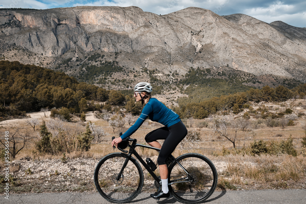 With a view of the beautiful mountains, a woman cyclist wearing a cycling kit and helmet is riding a gravel bike. Alicante region in Spain.