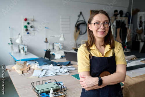 Successful smiling female tailor keeping her arms crossed on chest while standing against workplace with supplies and looking at camera