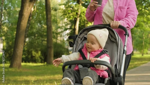 A cheerful little girl in a baby carriage, a young mother drinks coffee while walking with her child in an outdoor park