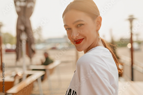 Outdoor photo of incredible stylish woman with dark hair and red lipstick posing to camera with happy smile in sunlight on blurred background in the city. 