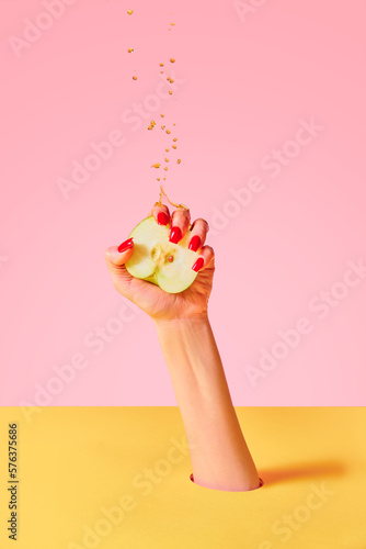 Tableau sur toile Closeup female hand with red painted nails squeezing half of apple fruit over pink yellow background