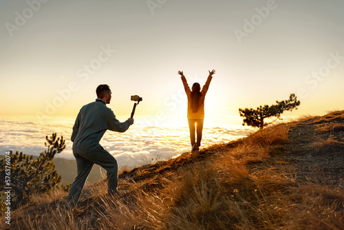Man tourist with electronic stabilizer gimbal is taking video shoot with happy girl at sunrise mountain top photo