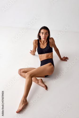 Top view of beautiful young brunette girl with slim fit body posing in black underwear over grey studio background. Concept of natural beauty, femininity, wellness, body care and fitness