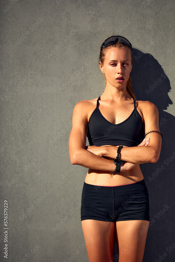 Obraz premium Shot of a flexible young woman warming up before running around the stadium, wearing sportswear, showing her stomach and abs, posing against a gray concrete wall