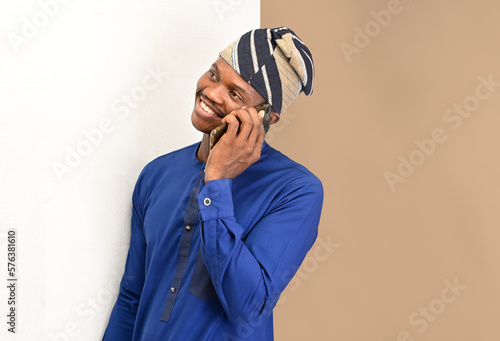 Young African man in native clothes calls on the phone on an isolatable background, stands in a splitted backdrop , communicates on a smartphone and looks away,  photo