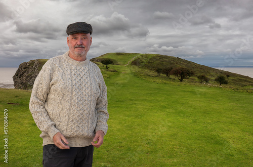 An elderly man wears a stylish Irish sheep's wool sweater and tweet cap. He on a green grassy area near the shore.