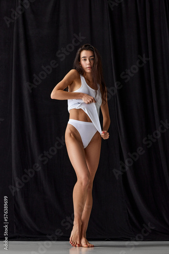 Beautiful young brunette girl with slim body posing in underwear, home wear over black studio background. Concept of natural beauty, tenderness, comfort, femininity, body care and fitness