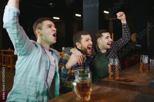 Soccer fans cheering for favourite team while enjoying drinks. Group of supporters celebrating victory while watching football match on tv in a club. Happy friends shouting while cheering for a goal.