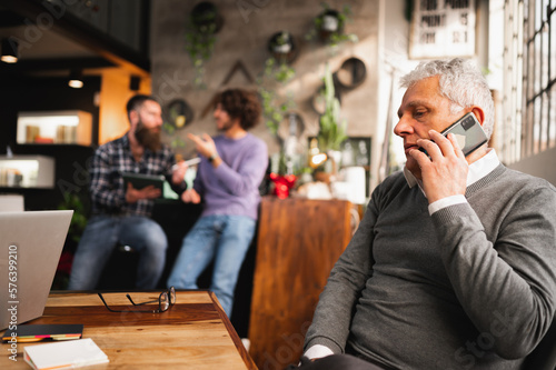 senior businessman talking on the phone sitting at desk in a modern office while colleagues discussing on background.