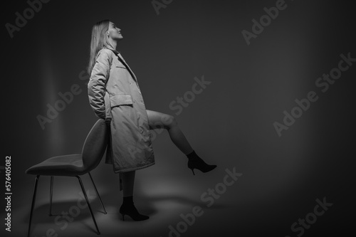 space, young, beautiful, emotional girl in a dark raincoat stands near a chair, raised one leg, leaning on a white background, a ray of light. Low key, ray of light