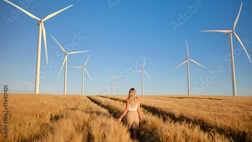Young blonde woman walks through a golden wheat field with wind turbines on the horizon and dreams of a clean and sustainable future for humanity