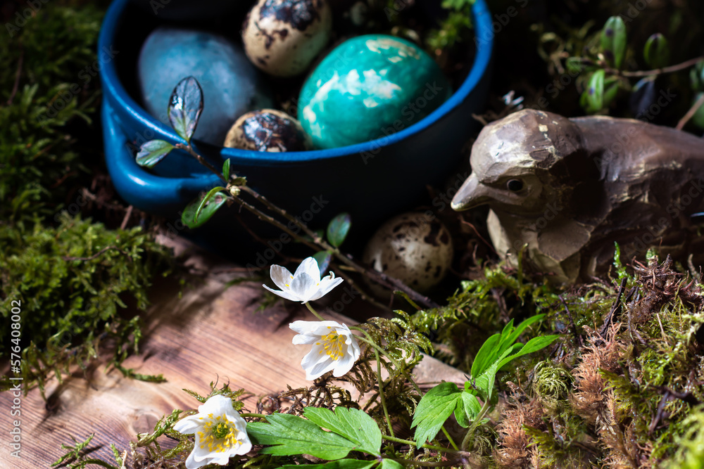 Blossom of windflower (anemone) in background of colored Easter eggs in blue dish, branches with buds, moss and wooden bird on wooden background, Easter eggs, Easter table decoration Blossom of windflower (anemone) in background of colored Easter eggs in blue dish, branches with buds, moss and wooden bird on wooden background, Easter eggs, Easter table decoration