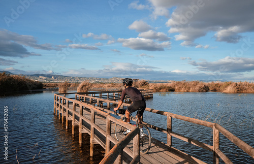 A man is riding over a lake with a background of mountains. Male cyclist is riding a gravel bike on a wooden bridge over a lake. Elche, Alicante, Spain