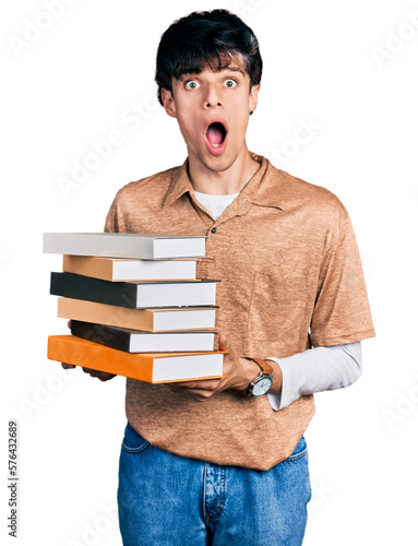 Handsome hipster young man holding a pile of books afraid and shocked with surprise and amazed expression, fear and excited face.