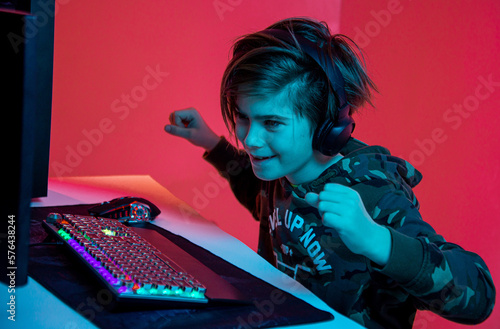 Excited boy looking at the screen of a gaming computer sitting in a cosy chair at home. Neon light in the room. Playing computer video games at home. Win a video game tournament. Self isolation © stenkovlad