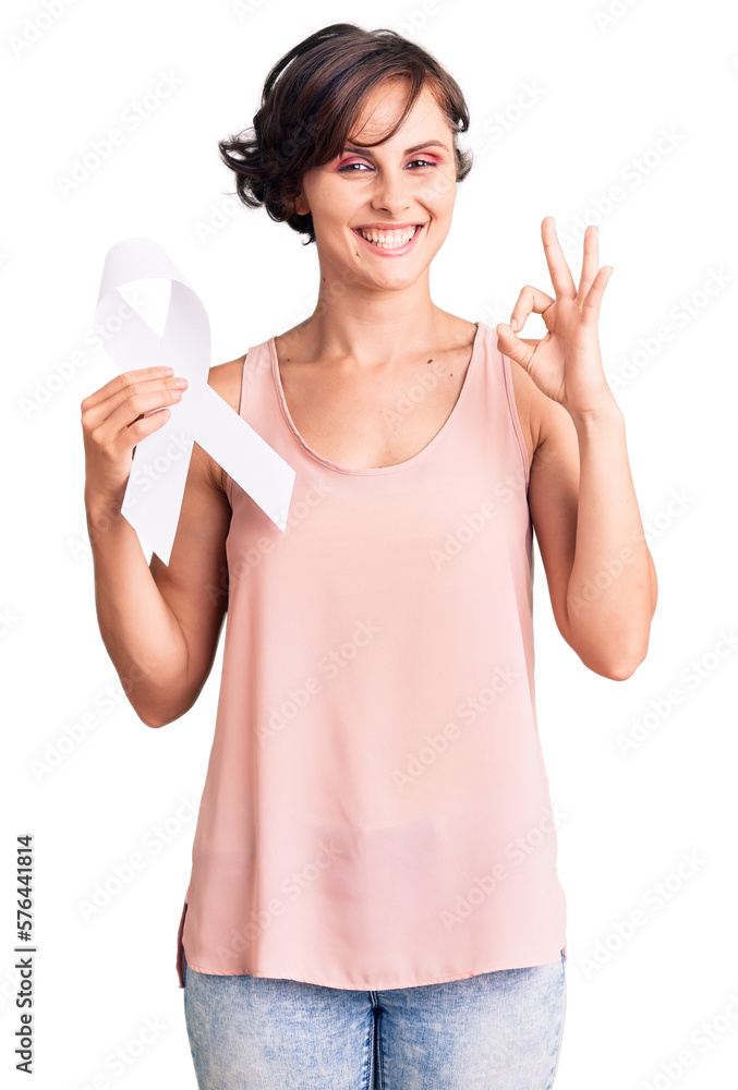 Beautiful young woman with short hair holding cancer awareness white ribbon doing ok sign with fingers, smiling friendly gesturing excellent symbol Beautiful young woman with short hair holding cancer awareness white ribbon doing ok sign with fingers, smiling friendly gesturing excellent symbol