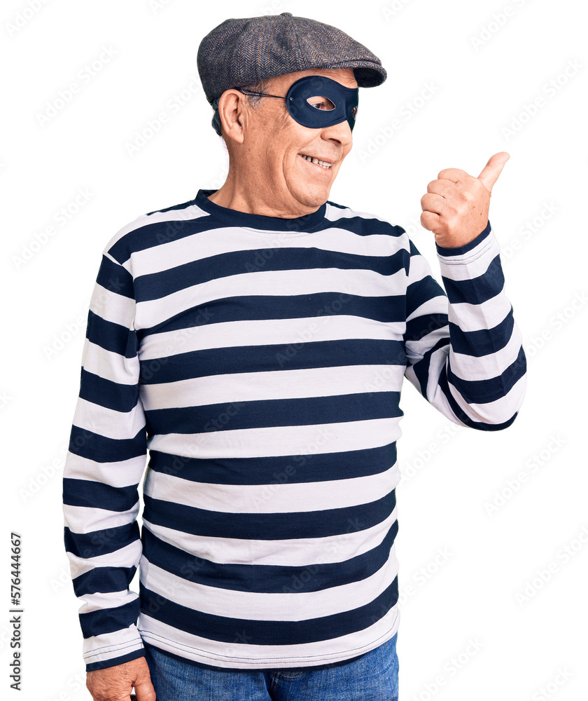 Senior handsome man wearing burglar mask and t-shirt smiling with happy face looking and pointing to the side with thumb up. Senior handsome man wearing burglar mask and t-shirt smiling with happy face looking and pointing to the side with thumb up.