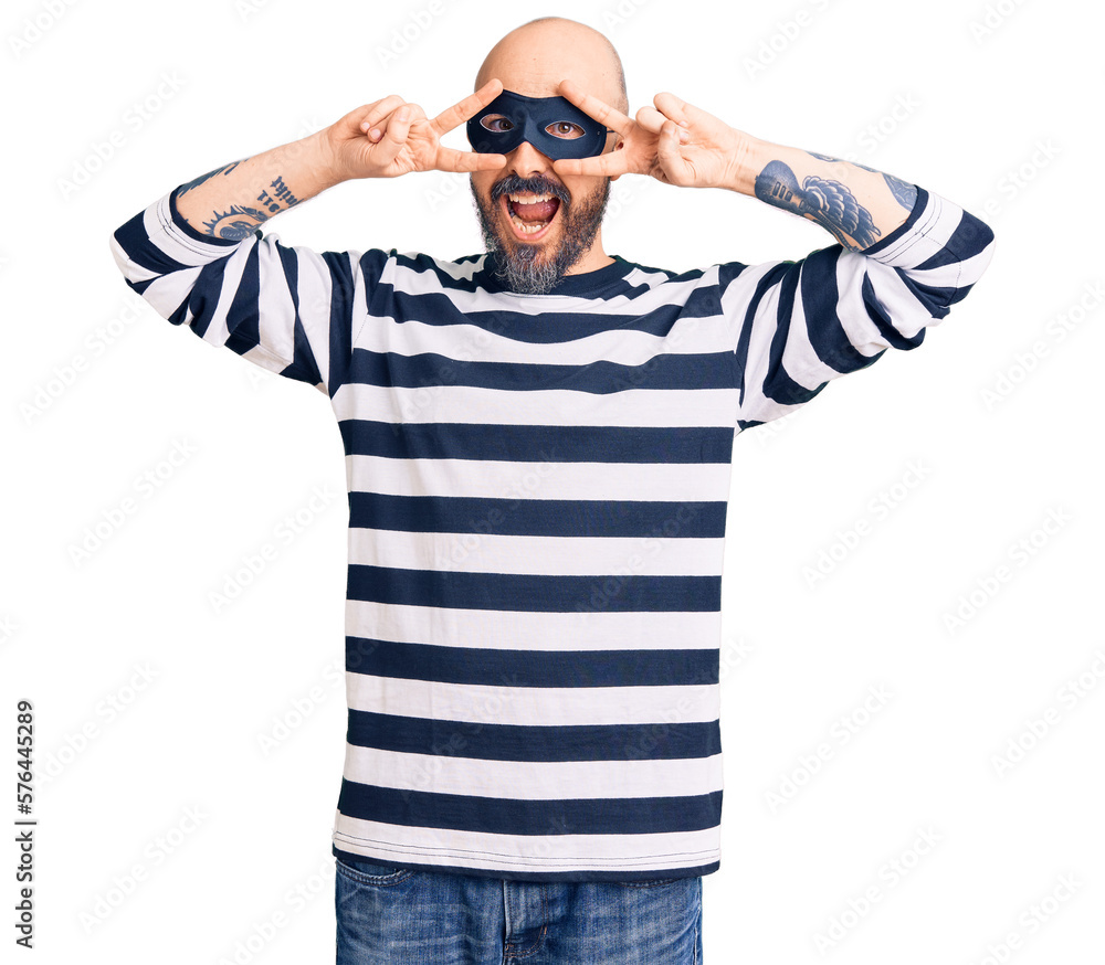 Young handsome man wearing burglar mask doing peace symbol with fingers over face, smiling cheerful showing victory Young handsome man wearing burglar mask doing peace symbol with fingers over face, smiling cheerful showing victory