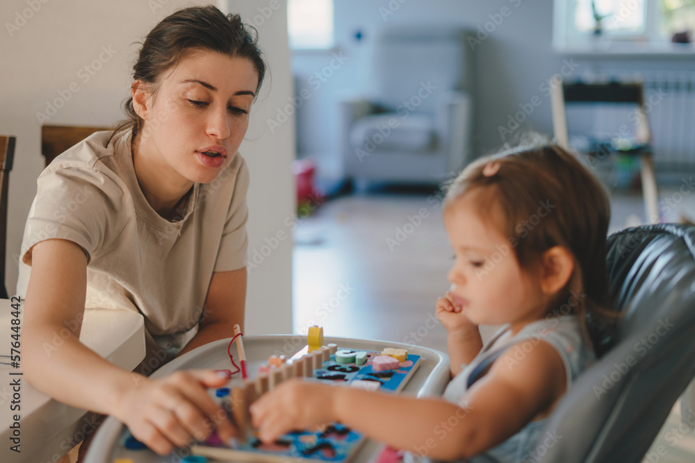 Fototapeta premium Attentive young mother involve baby girl in interesting game with colorful wooden components toy. Education concept. Family activity concept. Happy family kid