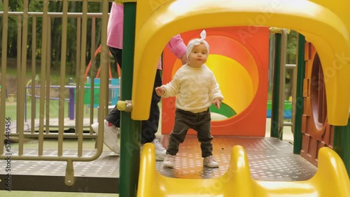 Young mother helping her baby girl with a bow on her head making first steps, little child with mom on the playground