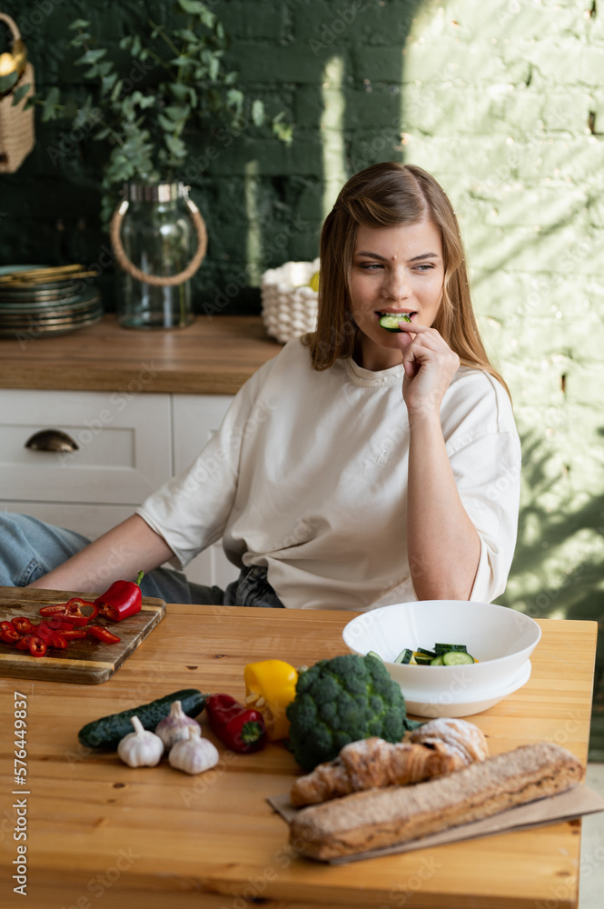 Photo on the topic of healthy eating. A young happy woman in the kitchen is sitting at a wooden table on which various vegetables and is eating a piece of cucumber while looking forward.