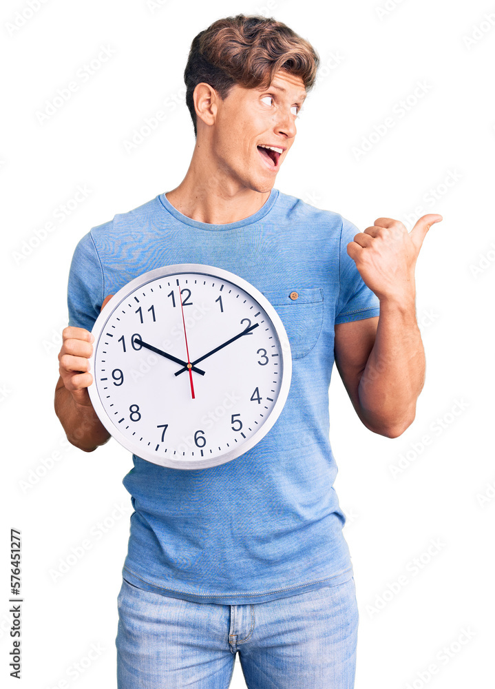 Young handsome man holding big clock pointing thumb up to the side smiling happy with open mouth Young handsome man holding big clock pointing thumb up to the side smiling happy with open mouth