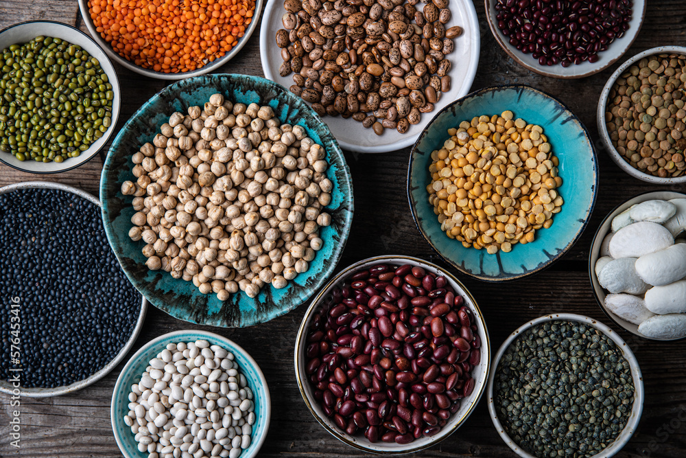 Different legumes (lentils, beans, chickpeas, mung, peas) in bowls and plates on wooden background.