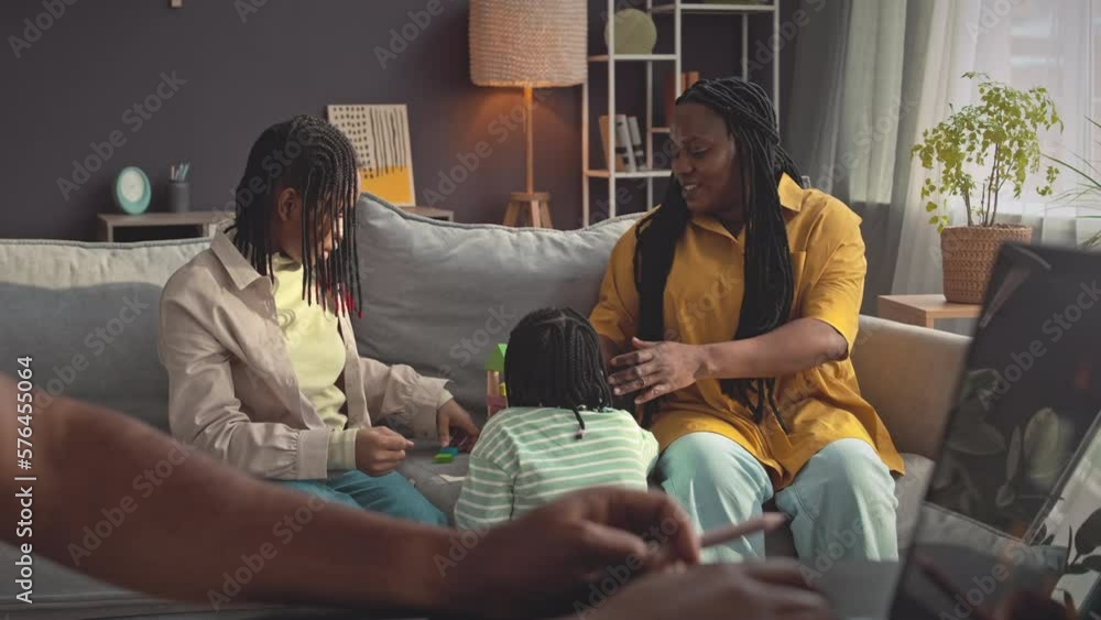 Black woman playing with two little daughters on couch in bright living room while her husband working on laptop from home Black woman playing with two little daughters on couch in bright living room while her husband working on laptop from home