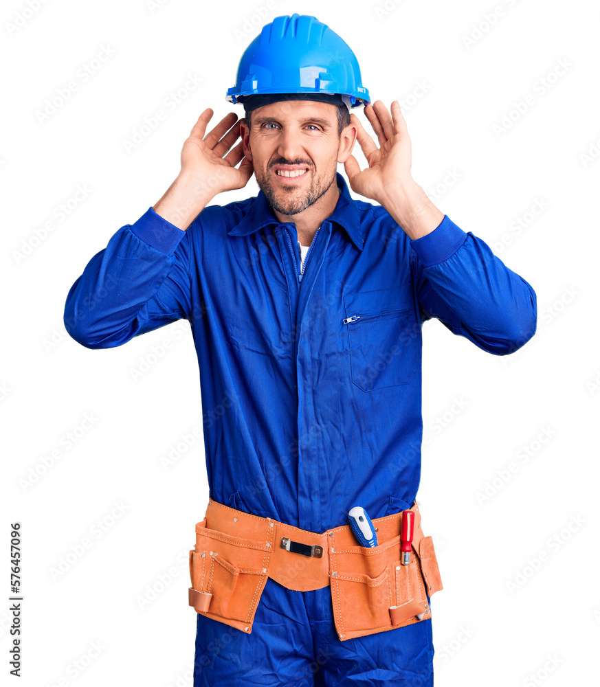 Young handsome man wearing worker uniform and hardhat trying to hear both hands on ear gesture, curious for gossip. hearing problem, deaf Young handsome man wearing worker uniform and hardhat trying to hear both hands on ear gesture, curious for gossip. hearing problem, deaf