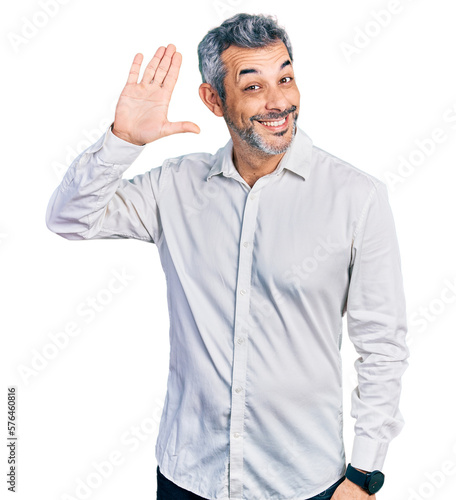 Middle age hispanic with grey hair wearing casual white shirt waiving saying hello happy and smiling, friendly welcome gesture