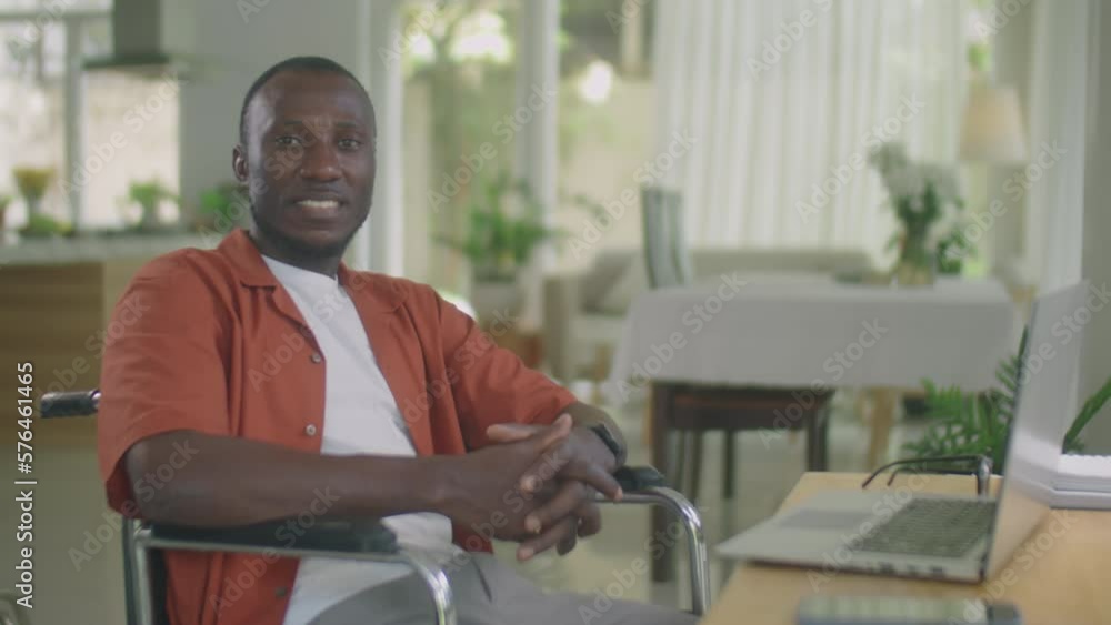 Medium portrait shot of African American man sitting in wheelchair at desk in home office and posing for camera with smile Medium portrait shot of African American man sitting in wheelchair at desk in home office and posing for camera with smile