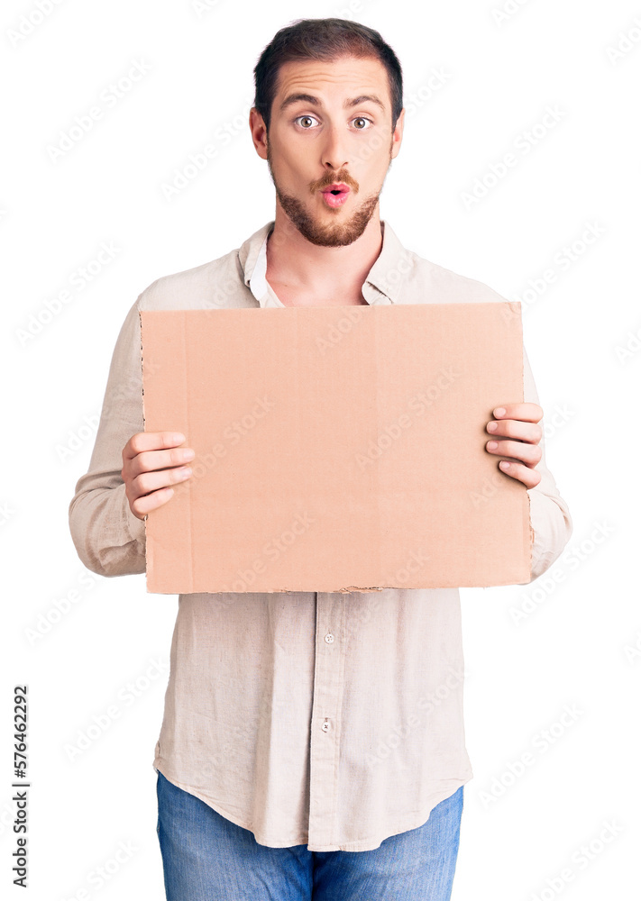 Young handsome caucasian man holding empty cardboard banner scared and amazed with open mouth for surprise, disbelief face Young handsome caucasian man holding empty cardboard banner scared and amazed with open mouth for surprise, disbelief face