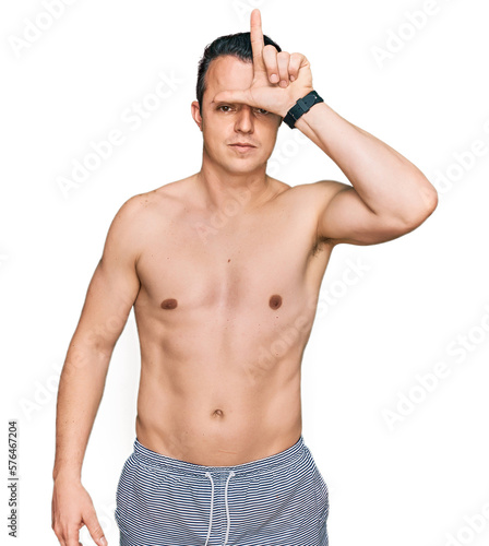 Handsome young man wearing swimwear shirtless making fun of people with fingers on forehead doing loser gesture mocking and insulting.