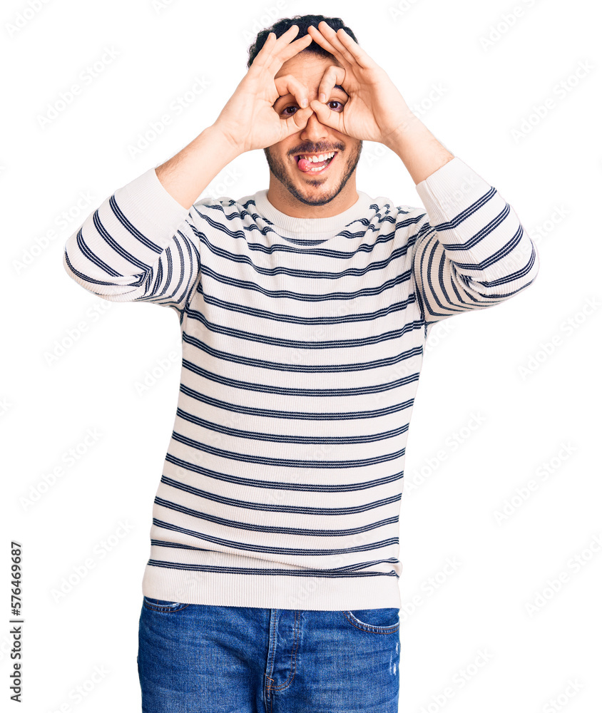 Young hispanic man wearing casual clothes doing ok gesture like binoculars sticking tongue out, eyes looking through fingers. crazy expression. Young hispanic man wearing casual clothes doing ok gesture like binoculars sticking tongue out, eyes looking through fingers. crazy expression.