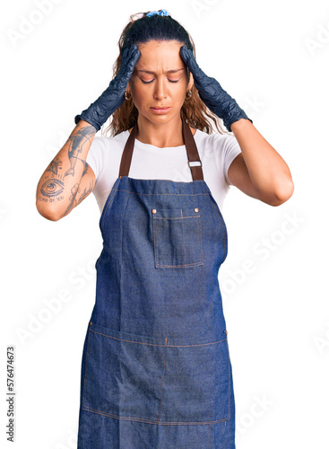 Obraz na plátně Young hispanic woman with tattoo wearing barber apron and gloves suffering from headache desperate and stressed because pain and migraine