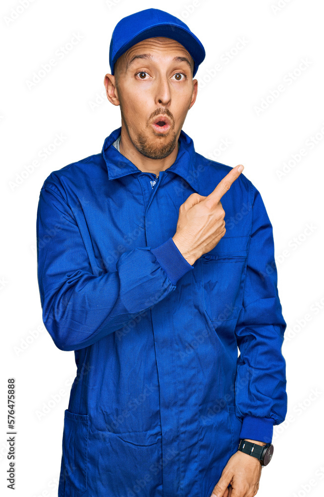 Bald man with beard wearing builder jumpsuit uniform surprised pointing with finger to the side, open mouth amazed expression.