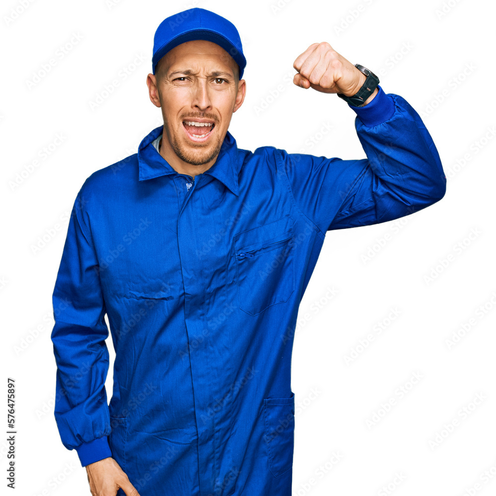 Bald man with beard wearing builder jumpsuit uniform angry and mad raising fist frustrated and furious while shouting with anger. rage and aggressive concept.