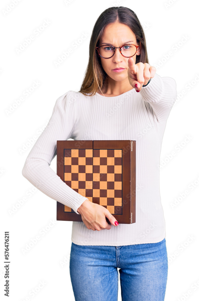 Beautiful brunette young woman holding chess pointing with finger to the camera and to you, confident gesture looking serious Beautiful brunette young woman holding chess pointing with finger to the camera and to you, confident gesture looking serious