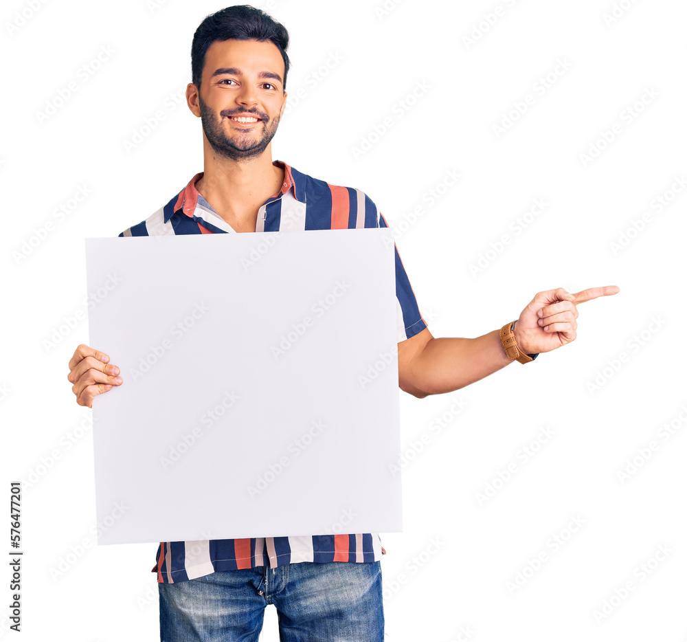Young handsome hispanic man holding cardboard banner with blank space smiling happy pointing with hand and finger to the side Young handsome hispanic man holding cardboard banner with blank space smiling happy pointing with hand and finger to the side