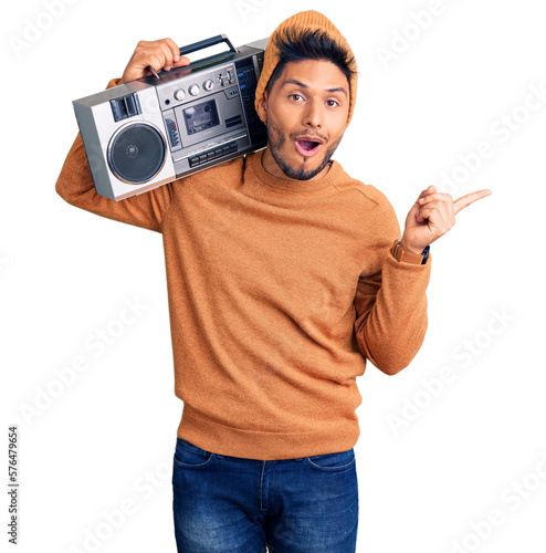 Handsome latin american young man holding boombox, listening to music surprised pointing with finger to the side, open mouth amazed expression.