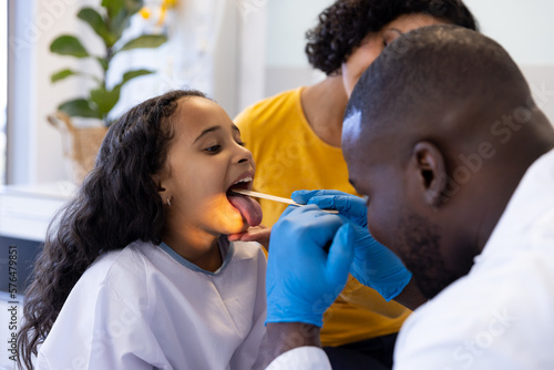Happy diverse male doctor examining throat of girl with mother in hospital